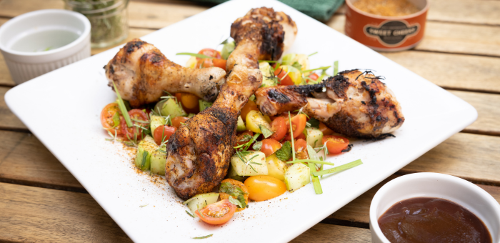 Char-grilled chicken drumsticks with caramelized skin stacked sitting on a bed of cucumber and tomato salad on a white plate and sitting on a wooden cutting board.
