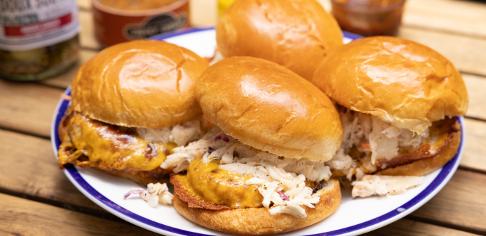 Barbecue-style chicken burgers with cheddar cheese and creamy slaw on toasted brioche buns, served on a white and blue plate with pickles and condiments in the background.
