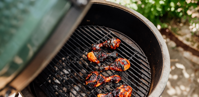 Barbecue chicken drumsticks with crispy charred skin cooking on an open round grill with glowing charcoal beneath.