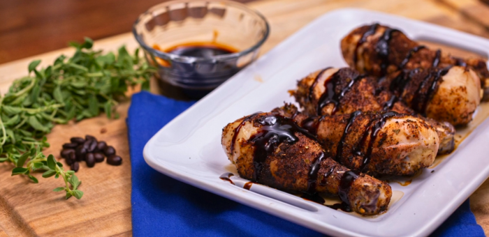Coffee-rubbed chicken drumsticks drizzled with balsamic glaze on a rectangular white plate, with herbs, coffee beans, and a sauce bowl in the background.