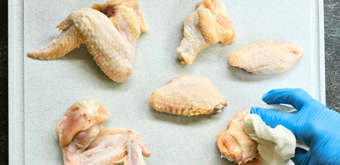 Raw chicken wings laid out on a tray being patted dry with a paper towel by a gloved hand before seasoning for the grill.