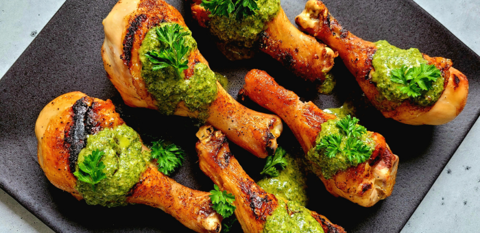 Grilled chicken drumsticks topped with vibrant green chimichurri sauce and parsley, arranged on a black serving plate.