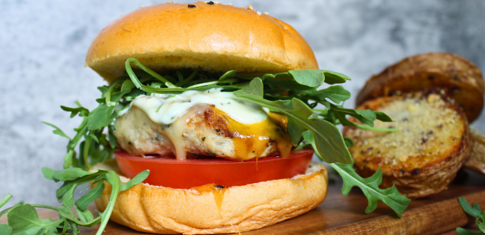 Gourmet grilled chicken burger with tomato, arugula, cheddar cheese, and herb mayo on a golden bun, served with roasted potatoes on a wooden board.