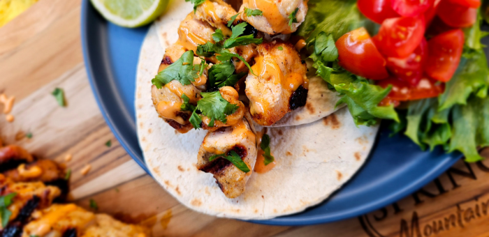 Close-up of grilled Bang Bang chicken tacos on a flour tortilla, topped with creamy spicy sauce and fresh cilantro, served with lettuce, tomatoes, and a lime wedge.
