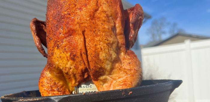 Seasoned whole chicken standing upright on a beer can in a cast iron pan, cooked outdoors with golden crispy skin, ready for serving as classic beer can chicken.
