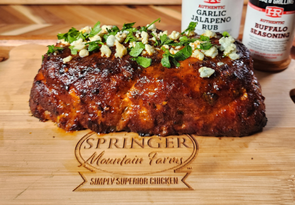 Buffalo chicken meatloaf garnished with blue cheese crumbles and chopped parsley, resting on a Springer Mountain Farms branded wooden cutting board with Heath Riles Garlic Jalapeno Rub and Buffalo Seasoning jars in the background.