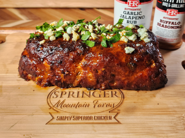 Buffalo chicken meatloaf garnished with blue cheese crumbles and chopped parsley, resting on a Springer Mountain Farms branded wooden cutting board with Heath Riles Garlic Jalapeno Rub and Buffalo Seasoning jars in the background.