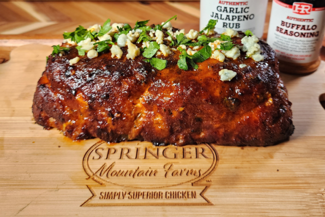 Buffalo chicken meatloaf garnished with blue cheese crumbles and chopped parsley, resting on a Springer Mountain Farms branded wooden cutting board with Heath Riles Garlic Jalapeno Rub and Buffalo Seasoning jars in the background.