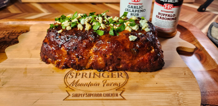 Buffalo chicken meatloaf glazed with sauce and topped with chopped herbs and blue cheese crumbles, served on a Springer Mountain Farms branded cutting board with garlic jalapeño and buffalo seasoning bottles in the background.