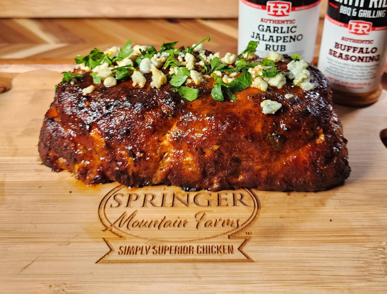 Buffalo chicken meatloaf garnished with blue cheese crumbles and chopped parsley, resting on a Springer Mountain Farms branded wooden cutting board with Heath Riles Garlic Jalapeno Rub and Buffalo Seasoning jars in the background.