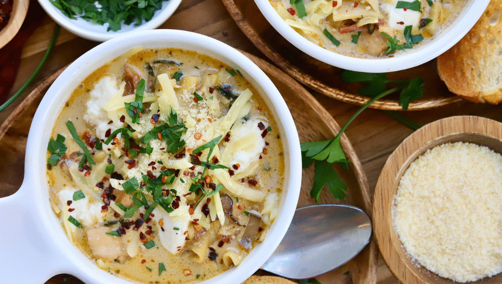Bowl of cheesy white lasagna soup with Springer Mountain Farms chicken, topped with melted cheese, fresh parsley, and red pepper flakes, served with Parmesan and toasted bread.