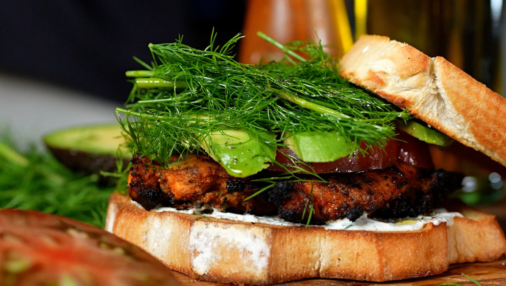 Grilled chicken bacon ranch sandwich stacked with juicy chicken breast, crispy bacon, avocado, fresh dill, heirloom tomato, and creamy ranch dressing on toasted white bread.