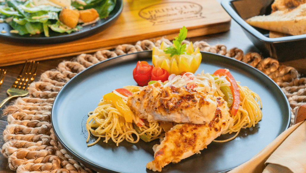 Creamy Chicken Scampi served over angel hair pasta with sautéed bell peppers, garnished with carved vegetables on a black dinner plate, paired with salad and toasted bread in the background on a rustic table.