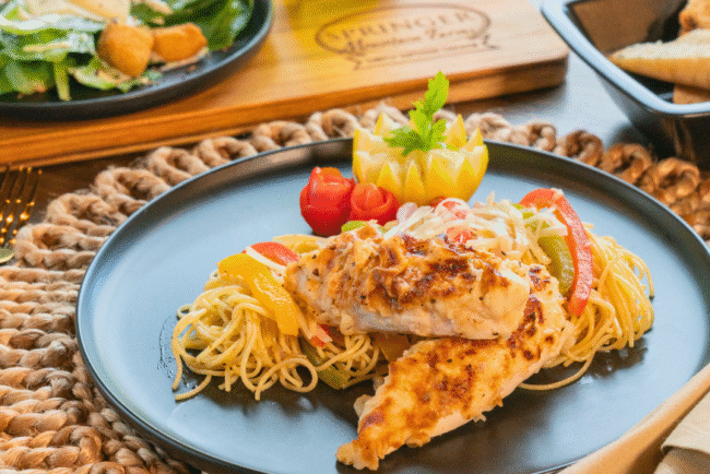 Creamy Chicken Scampi served over angel hair pasta with sautéed bell peppers, garnished with carved vegetables on a black dinner plate, paired with salad and toasted bread in the background on a rustic table.