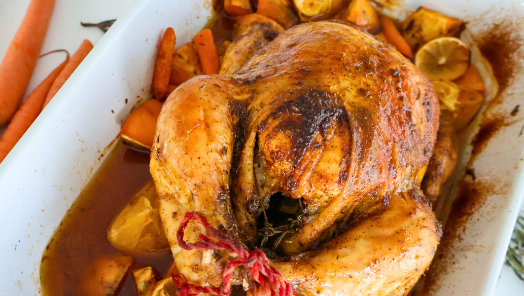 Golden brown citrus herb roasted whole Springer Mountain Farms chicken resting on carrots, sweet potatoes, lemons, and fresh herbs in a white baking dish, showcasing crispy skin and juicy pan drippings.