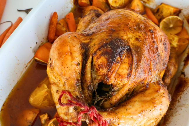 Golden brown citrus herb roasted whole Springer Mountain Farms chicken resting on carrots, sweet potatoes, lemons, and fresh herbs in a white baking dish, showcasing crispy skin and juicy pan drippings.