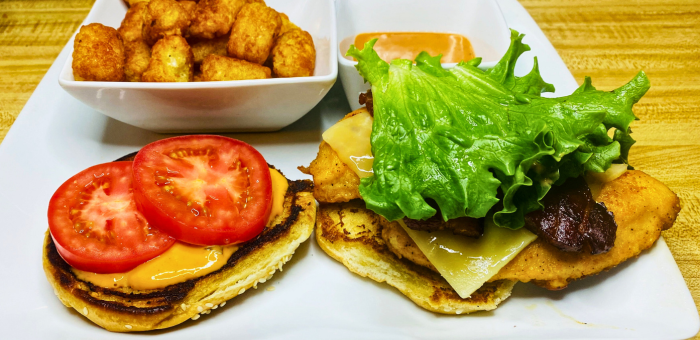 Crispy fried chicken sandwich with lettuce, tomato, cheese, and bacon on a toasted bun, served with golden tater tots and dipping sauce.