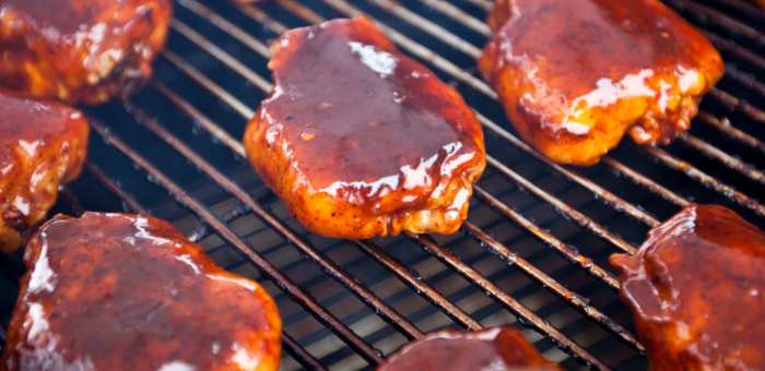 Close-up view of bone-in, skin-on chicken thighs sizzling on a grill, coated in a glossy layer of barbecue sauce. The rich, caramelized glaze gives the chicken a mouthwatering finish as smoke rises between the grill grates.