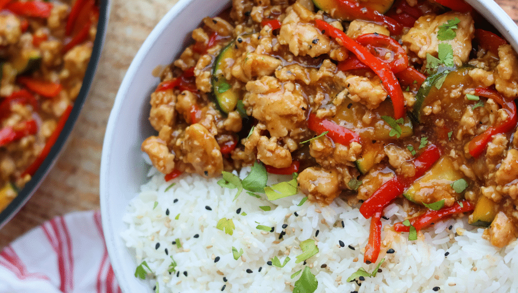 Savory ground chicken stir fry with red bell peppers, zucchini, and a flavorful sauce served over fluffy white rice with sesame seeds and fresh herbs.