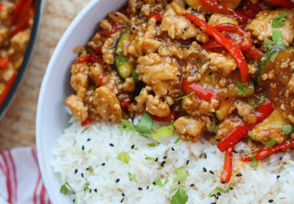 Savory ground chicken stir fry with red bell peppers, zucchini, and a flavorful sauce served over fluffy white rice with sesame seeds and fresh herbs.