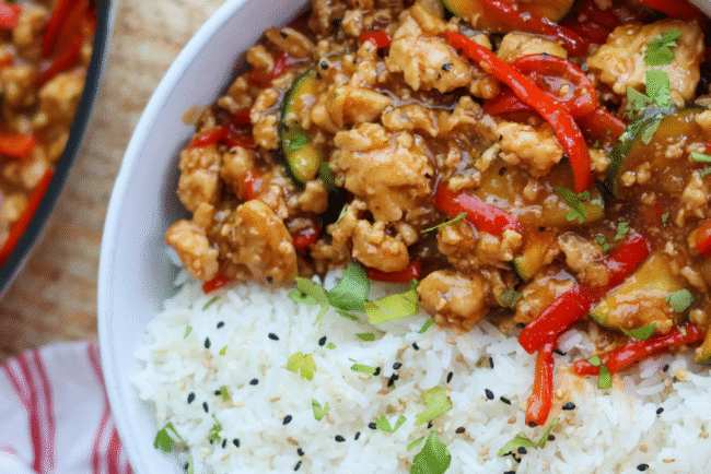 Savory ground chicken stir fry with red bell peppers, zucchini, and a flavorful sauce served over fluffy white rice with sesame seeds and fresh herbs.