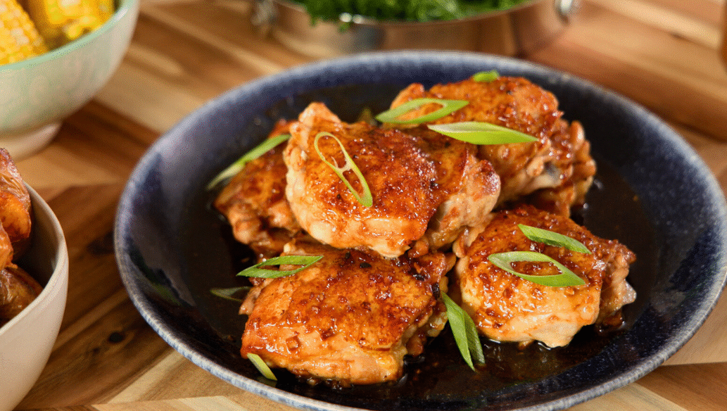 Juicy honey garlic glazed chicken thighs topped with sliced green onions on a dark plate, served with roasted potatoes and corn for a cozy homemade dinner.