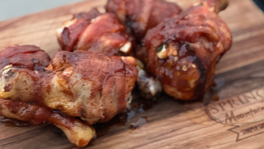Grilled jalapeño popper stuffed chicken drumsticks wrapped in crispy bacon, glazed with barbecue sauce, served on a Springer Mountain Farms wooden board.