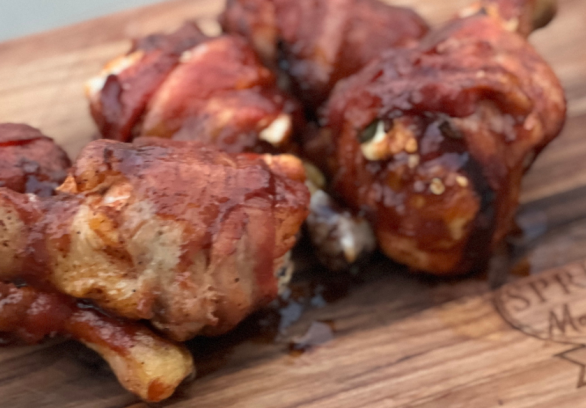 Grilled jalapeño popper stuffed chicken drumsticks wrapped in crispy bacon, glazed with barbecue sauce, served on a Springer Mountain Farms wooden board.