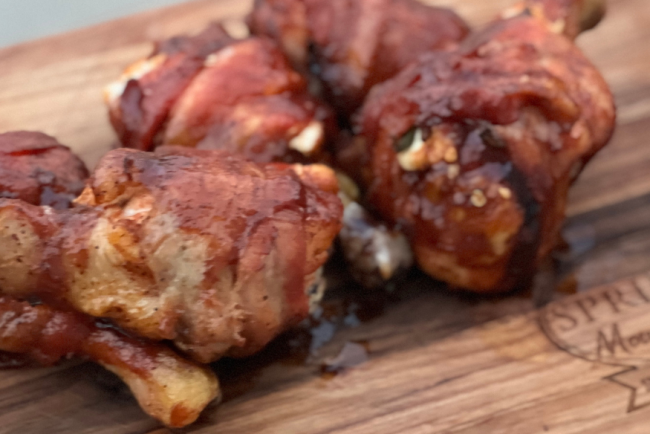 Grilled jalapeño popper stuffed chicken drumsticks wrapped in crispy bacon, glazed with barbecue sauce, served on a Springer Mountain Farms wooden board.