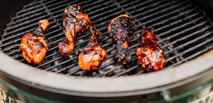 Korean BBQ chicken drumsticks cooking on a grill, charred and caramelized to perfection with a glossy, deep-red glaze. The drumsticks are arranged over open grates inside a ceramic grill, showcasing a rich, smoky finish and bold flavors.