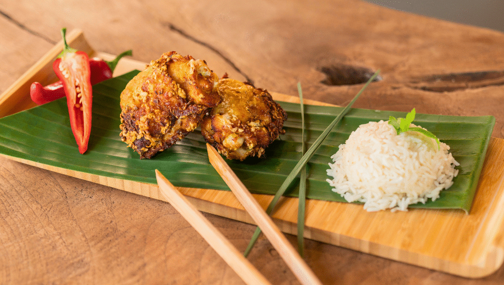 Malaysian Fried Chicken Thighs (Ayam Goreng) served on a banana leaf-lined wooden tray with crispy golden chicken pieces, sliced red chili, a mound of steamed white rice, and chopsticks, styled on a rustic wooden table.