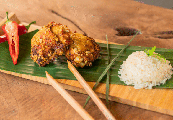 Malaysian Fried Chicken Thighs (Ayam Goreng) served on a banana leaf-lined wooden tray with crispy golden chicken pieces, sliced red chili, a mound of steamed white rice, and chopsticks, styled on a rustic wooden table.