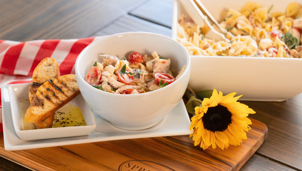 Marry Me Chicken Pasta Salad served in a white bowl with cherry tomatoes, fresh herbs, and tender chicken, accompanied by grilled baguette slices and olive oil dip on a Springer Mountain Farms wooden cutting board, set on a rustic table with a red gingham napkin and a sunflower for décor.