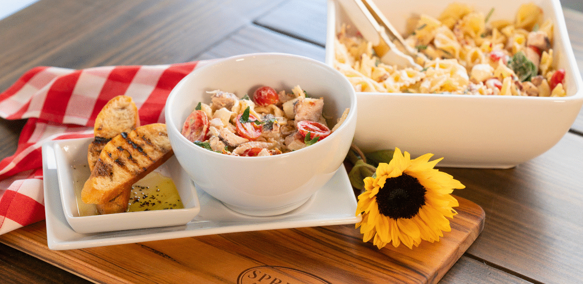 Marry Me Chicken Pasta Salad served in a white bowl with cherry tomatoes, fresh herbs, and tender chicken, accompanied by grilled baguette slices and olive oil dip on a Springer Mountain Farms wooden cutting board, set on a rustic table with a red gingham napkin and a sunflower for décor.