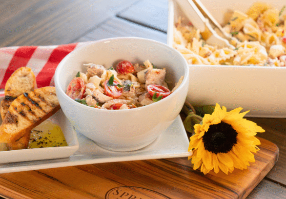 Marry Me Chicken Pasta Salad served in a white bowl with cherry tomatoes, fresh herbs, and tender chicken, accompanied by grilled baguette slices and olive oil dip on a Springer Mountain Farms wooden cutting board, set on a rustic table with a red gingham napkin and a sunflower for décor.