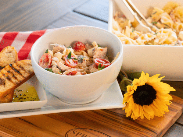 Marry Me Chicken Pasta Salad served in a white bowl with cherry tomatoes, fresh herbs, and tender chicken, accompanied by grilled baguette slices and olive oil dip on a Springer Mountain Farms wooden cutting board, set on a rustic table with a red gingham napkin and a sunflower for décor.
