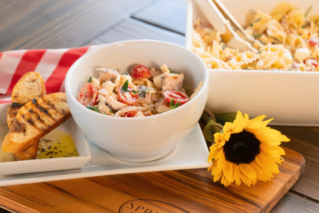 Marry Me Chicken Pasta Salad served in a white bowl with cherry tomatoes, fresh herbs, and tender chicken, accompanied by grilled baguette slices and olive oil dip on a Springer Mountain Farms wooden cutting board, set on a rustic table with a red gingham napkin and a sunflower for décor.