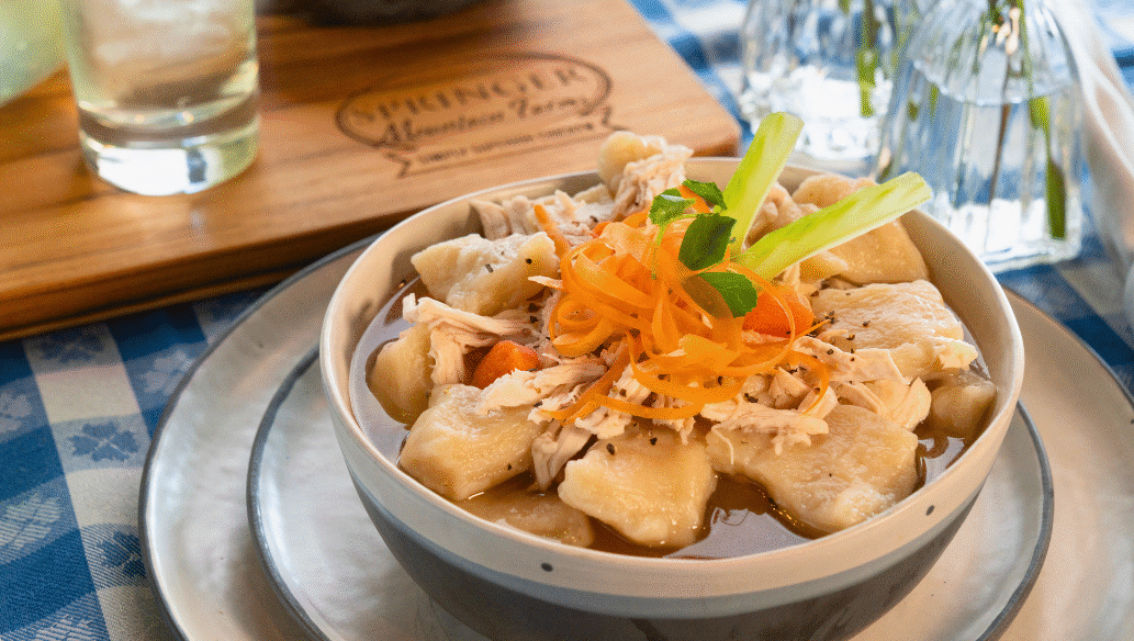 Bowl of old-fashioned Southern chicken and dumplings made with tender Springer Mountain Farms shredded chicken, fluffy dumplings, carrots, and rich broth, garnished with carrot ribbons and celery on a checkered tablecloth.