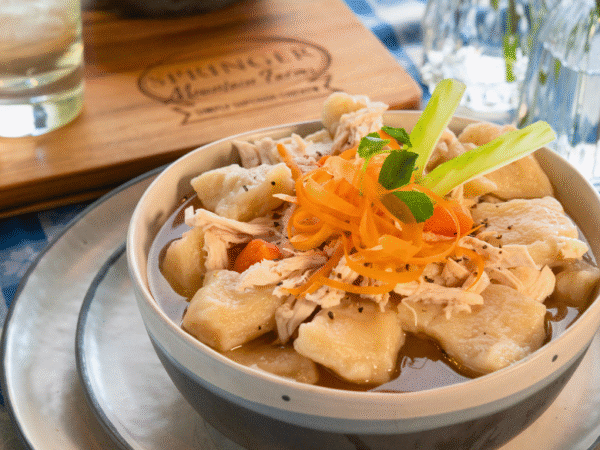 Bowl of old-fashioned Southern chicken and dumplings made with tender Springer Mountain Farms shredded chicken, fluffy dumplings, carrots, and rich broth, garnished with carrot ribbons and celery on a checkered tablecloth.