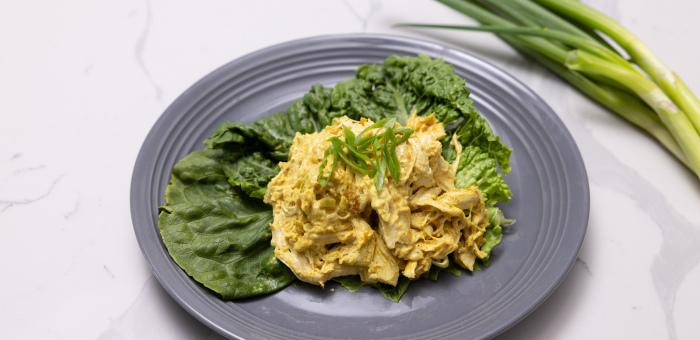 Shredded chicken coated in creamy yellow curry sauce, served on a bed of fresh green leaf lettuce on a gray plate with scallions on the side.
