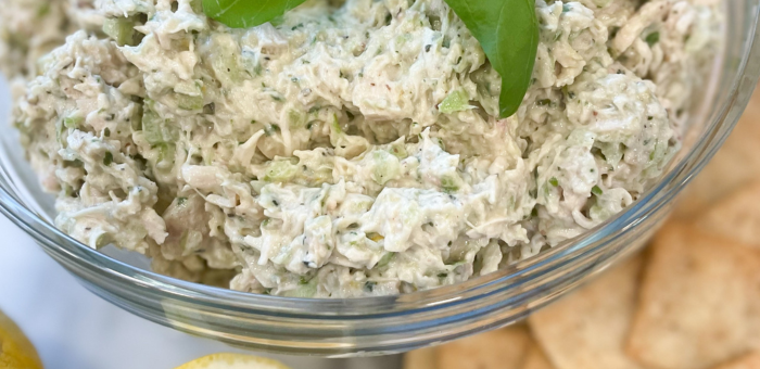 Close-up of a glass bowl filled with creamy pesto chicken salad made with shredded chicken, fresh basil, and diced celery, served with pita chips.