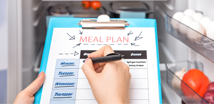 Person writing a weekly family meal plan on a clipboard in front of an open refrigerator with fresh ingredients.