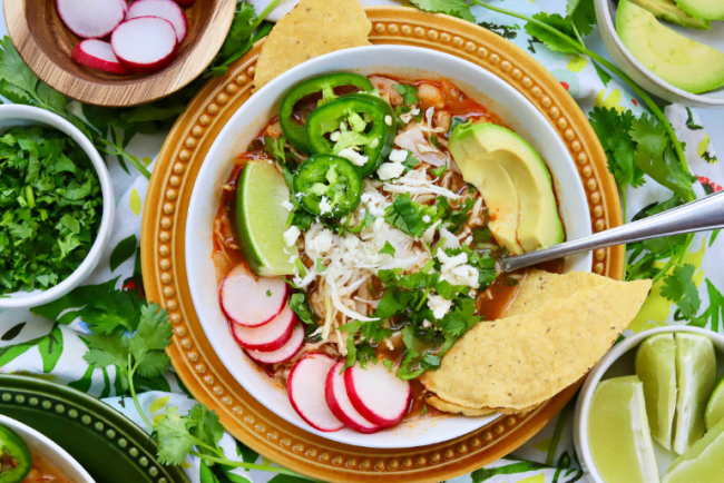 A bowl of pozole rojo with tender chicken, vibrant red broth, and hominy. The soup is garnished with fresh toppings, including sliced radishes, shredded cabbage, jalapeno slices, avocado, lime wedges, and a sprinkle of cotija cheese, creating a colorful and appetizing presentation.