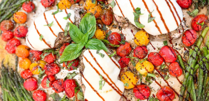 Sheet pan with baked chicken breasts topped with mozzarella, cherry tomatoes, asparagus, and fresh basil drizzled with balsamic glaze.