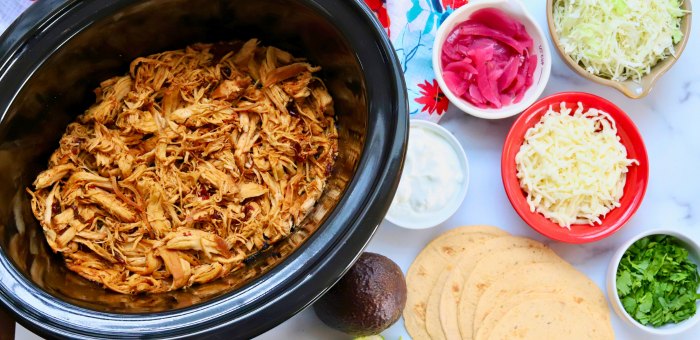 Slow cooker filled with shredded honey chipotle chicken surrounded by taco toppings and tortillas.