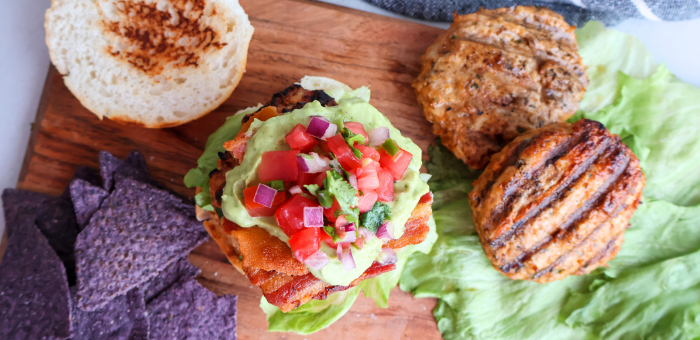 Grilled chicken burger topped with guacamole, pico de gallo, and crispy bacon on a toasted bun with lettuce, served alongside grilled patties and blue corn tortilla chips on a wooden board.
