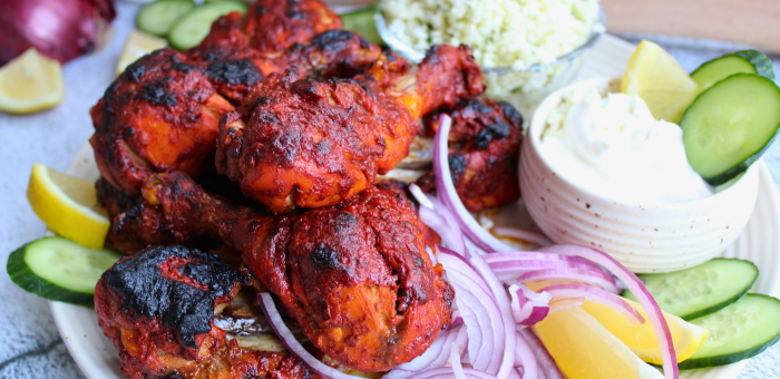 Charred, richly spiced Tandoori chicken drumsticks served with lemon wedges, sliced cucumber, red onion, and a bowl of creamy yogurt sauce.