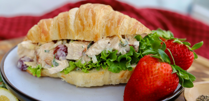 Creamy chicken salad with grapes, celery, and leafy greens served on a buttery croissant, plated with fresh strawberries on the side.