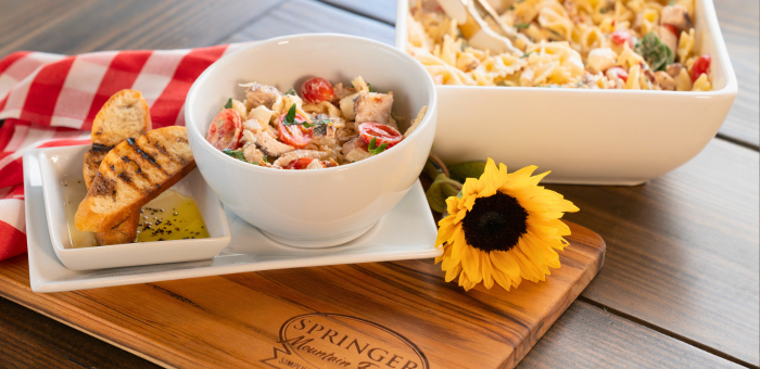 Marry Me Chicken Pasta Salad served in a white bowl with cherry tomatoes, fresh herbs, and tender chicken, accompanied by grilled baguette slices and olive oil dip on a Springer Mountain Farms wooden cutting board, set on a rustic table with a red gingham napkin and a sunflower for décor.