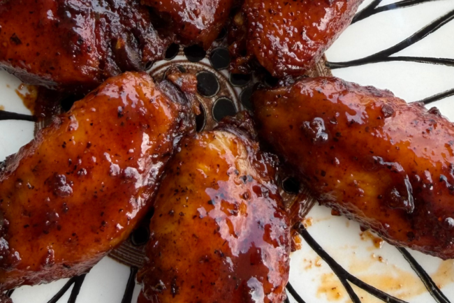 Close-up of smoky barbecue chicken wings coated in a thick, glossy BBQ sauce, arranged on a decorative plate with a rich caramelized finish and charred edges.